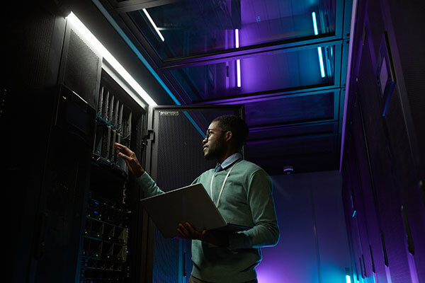 An African-American data engineer working on a supercomputer in a server room. Blue and purple neon lights add a dramatic, cyberpunk appearance to the racks.