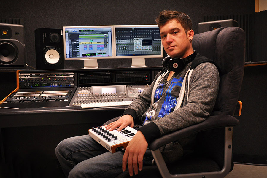 A USV electronic music production student working hands-on in the studio, a mixing keyboard on his lap and audio editing software on a dual monitors.